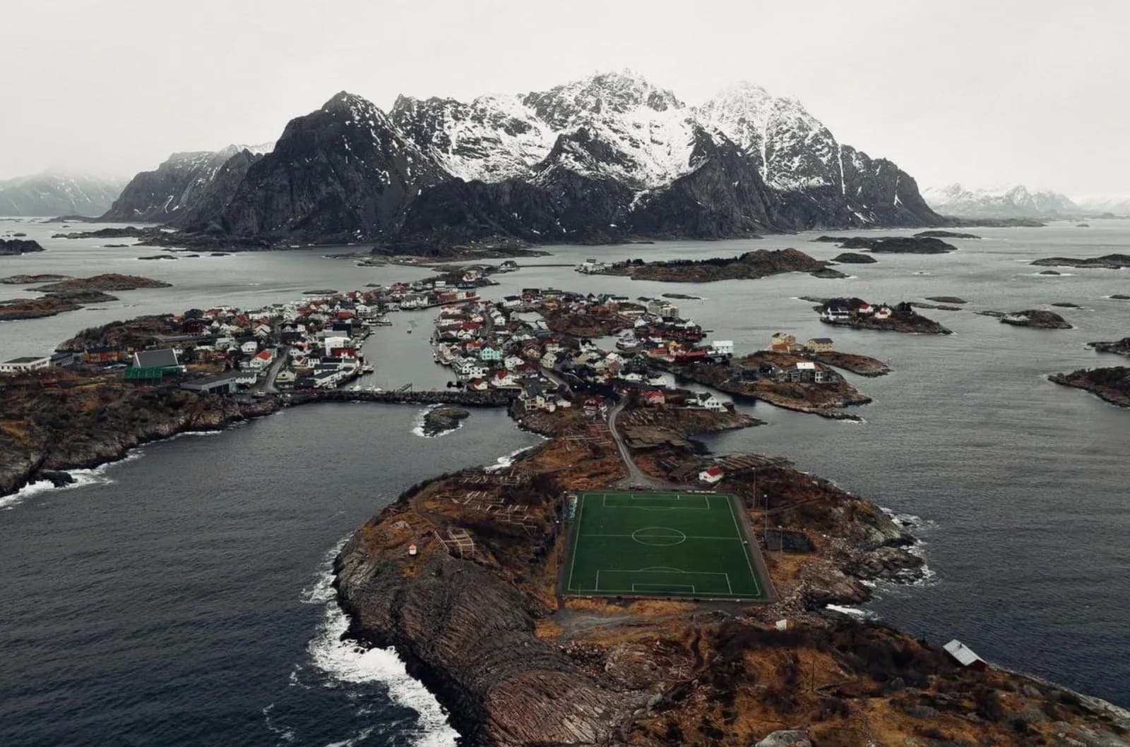Luftaufnahme der Lofoten: Fußballplatz auf einer Felsinsel, dahinter verschneite Berge